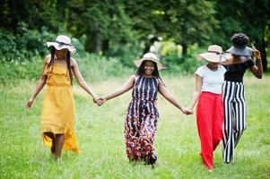 Group of four gorgeous african american womans wear summer hat spending time at green grass in park. photo