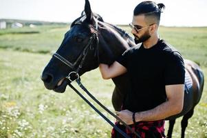 Arab tall beard man wear in black and sunglasses with arabian horse. photo