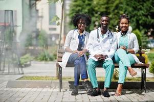 Three African American group doctors with stethoscope wearing lab coat sitting on bench. photo