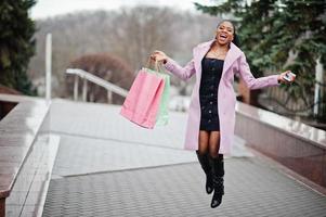 joven y elegante mujer afroamericana en la calle, vestida con un abrigo de moda con bolsas de compras y teléfono móvil en las manos. foto