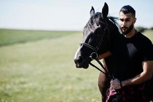 Hombre árabe de barba alta vestido de negro con caballo árabe. foto