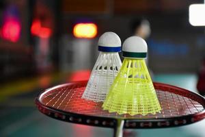 White and yellow plastic badminton shuttlecock and racket with neon light shading on green floor in indoor badminton court, blurred badminton background, copy space. photo