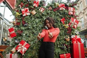 Portrait of a curly haired african woman wearing fashionable red turtleneck posing against christmas decorations, new year eve theme. Show thumbs up. photo