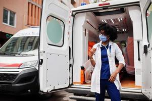 African american female paramedic in face protective medical mask standing in front of ambulance car. photo