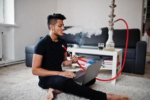 hombre indio guapo y de moda en negro sentado en la habitación, fumando narguile y trabajando en la computadora portátil. foto