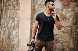 Impresionante y hermoso fotógrafo de hombre macho con barba árabe alta con gafas y camiseta negra con cámara profesional en las manos. foto