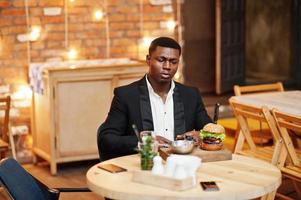 Respectable young african american man in black suit sitting in restaurant with tasty double burger and soda drink. photo