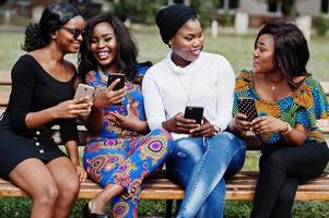 grupo de cuatro chicas afroamericanas sentadas en un banco al aire libre con teléfonos móviles en las manos. foto