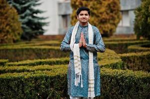 Indian man wear on traditional clothes with white scarf posed outdoor against green bushes at park, show namaste hands sign. photo