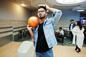 Stylish asian man in jeans jacket and glasses standing at bowling alley with ball at hand. photo