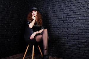 Brunette curly haired long legs girl in black leather dress and cap posed at studio on chair against dark brick wall. photo