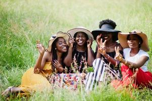 Group of four gorgeous african american womans wear summer hat sitting at green grass in park. photo