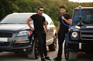 Two asian brothers man wear on all black posed near suv cars and smoke hookah. photo