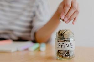 la mano de una mujer asiática está poniendo una moneda en una botella de vidrio sobre una mesa de madera, negocios de inversión, jubilación, finanzas y ahorro de dinero para el concepto futuro. foto