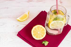 Mason jar glass of homemade lemonade with lemons, mint and red paper straw on coth napkin and wooden background. Summer refreshing beverage. photo