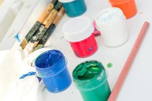 Artist's work place. Open multicolored gouache, paint brushes and pencil on white table. photo