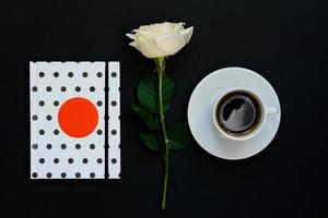 Cup of black coffee, notebook and white rose on black background, Top view, copy space. photo