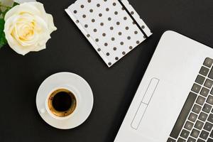 Flat lay, top view office table desk. Workspace with laptop, white rose, polka dot diary and coffee mug on black background. photo