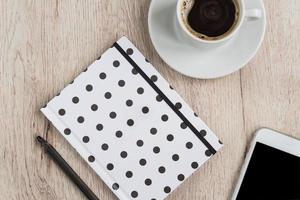 concepto de negocio y oficina: cuaderno con tapa de lunares en blanco y negro, teléfono inteligente y taza de café negro sobre una mesa de madera. vista superior. foto