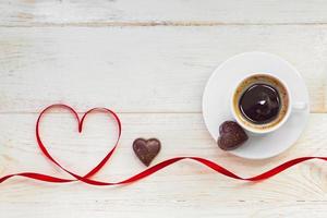 Cup of black americano coffe with gold plated chocolate in shape of heart and red satin ribbon on wooden background. Top view. photo