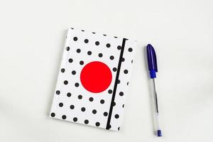 Black and white polka dot note book with red circle  on the cover and blue pen on white table. top view, minimal flat lay photo