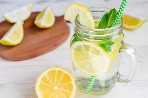 Mason jar glass of homemade lemonade with lemons, mint and green paper straw on wooden rustic background. Summer refreshing beverage. photo