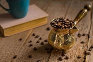 granos de café recién tostados en cezve cafetera turca tradicional libro cerrado y taza sobre mesa de madera. foto