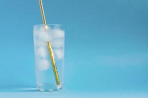 Close up of pure water with ice and gold paper straw in tall transparent glass and sun glare on blue background. Copy space photo