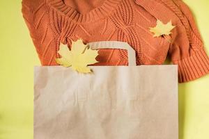 Orange woman warm sweater in paper craft package on yellow background with autumn maple leaves decoration. Clothing sale and black friday concept.  Top view. photo