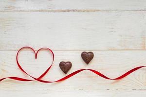 Red gift celebration ribbon waved in shape of heart and chocolate sweets on white wooden background. Greeting card with copy space for St Valentines Day photo