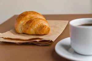 Tasty breackfast. French croissant served on craft paper and cup of black coffee or espresso on brown background. Copy space. photo