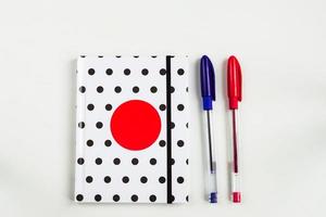 Black and white polka dot note book with red circle  on the cover and blue and red pens on white table. top view, minimal flat lay photo
