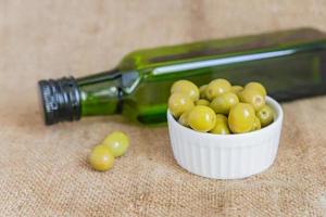 fresh marinated green olives in white ceramic bowl and green bottle of premium virgin olive oil on burlap cloth background photo