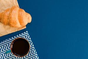 top view of cup of black coffe and croissant on craft paper on blue background with copy space. Morning breakfast in french style. photo