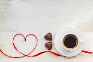 Cup of black americano coffe with gold plated chocolate in shape of heart and red satin ribbon on wooden background. Top view. Copy space. Valentines greeting card