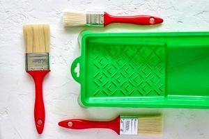 tres cepillos con asas rojas y bandeja de pintura verde sobre fondo de hormigón blanco. herramientas y accesorios para la renovación del hogar. vista superior foto