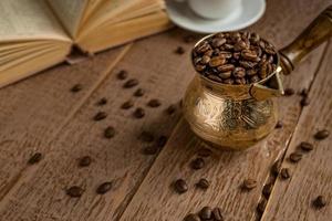granos de café recién tostados en cezve, cafetera tradicional turca, libro abierto y taza sobre una mesa de madera. foto