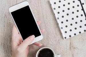 concepto de negocio y oficina - mano de mujer sosteniendo un smartwhone blanco junto al cuaderno de tapa de lunares en blanco y negro y una taza de café negro en la mesa de madera. vista superior. foto