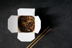 Asian restaurant food delivery. Soba noodles with meat, vegetables and soy sauce in white take-out paper box on black background with chopsticks, top view, copy space photo