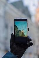 mujer da un guante de cuero negro con un smartphone negro para tomar una foto de la ciudad navideña en un día soleado