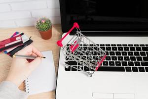 Small red shopping cart or trolley on laptop keyboard and woman hand writing in notes. Technology business online shopping concept photo