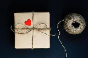 Handcrafted gift box wrapped in Craft paper with red wooden heart, rope and bow on black background. Top view, flat lay photo