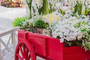 carretilla de madera roja con cajas de madera llenas de flores artificiales en flor. decoración de calles y jardines. foto