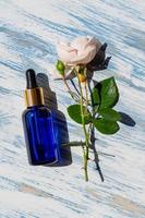 Cosmetic bottle dropper with garden rose flower and shadow on wooden blue background. Face and body care spa concept. Hyaluronic acid oil, serum with collagen and peptides skin care product. photo