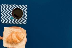 top view of cup of black coffe and croissant on a cutting board and craft paper on blue background with copy space. Morning breakfast in french style. photo