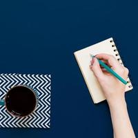 top view of cup of black coffe and woman hand whriting in notebook on blue background with copy space. photo
