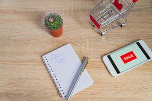 close up view of office desk with white smartphone with sale text, note and small shopping cart. Technology business online shopping concept photo