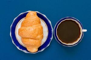 top view of cup of black coffe and croissant on a plate on blue background. Morning breakfast in french style. photo