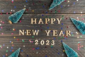 Happy New year 2023 celebration. Wooden text and numbers 2021 on wooden background with scattered multicolored confetti and fur tree miniatures. photo