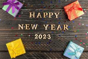 Multicolored gift boxes with tied bows. New year presents on wooden table with scattered confetti stars. Text Happy new year 2023. Festive greeting card. Flat lay. photo